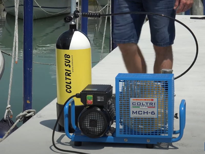 A man in short stands on the dock filling his diving cylinder with a blue Coltri high pressure air compressor.