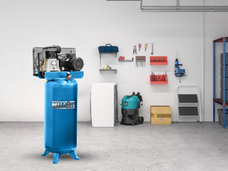 A blue Nuair belt driven vertical air compressor stands in a garage which has plain panted walls and other items such a a step ladder and wall mounted tools in the background
