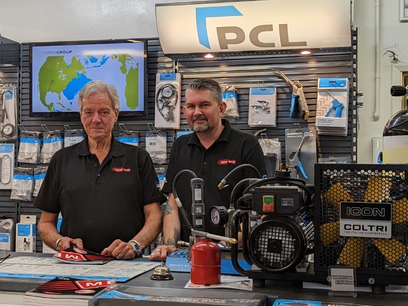Metro Sales owner Gary with Neil standing behind the shop counter with a high pressure Coltri air compressor in front of them. They are wearing black Metro Sales t-shirts and smiling.